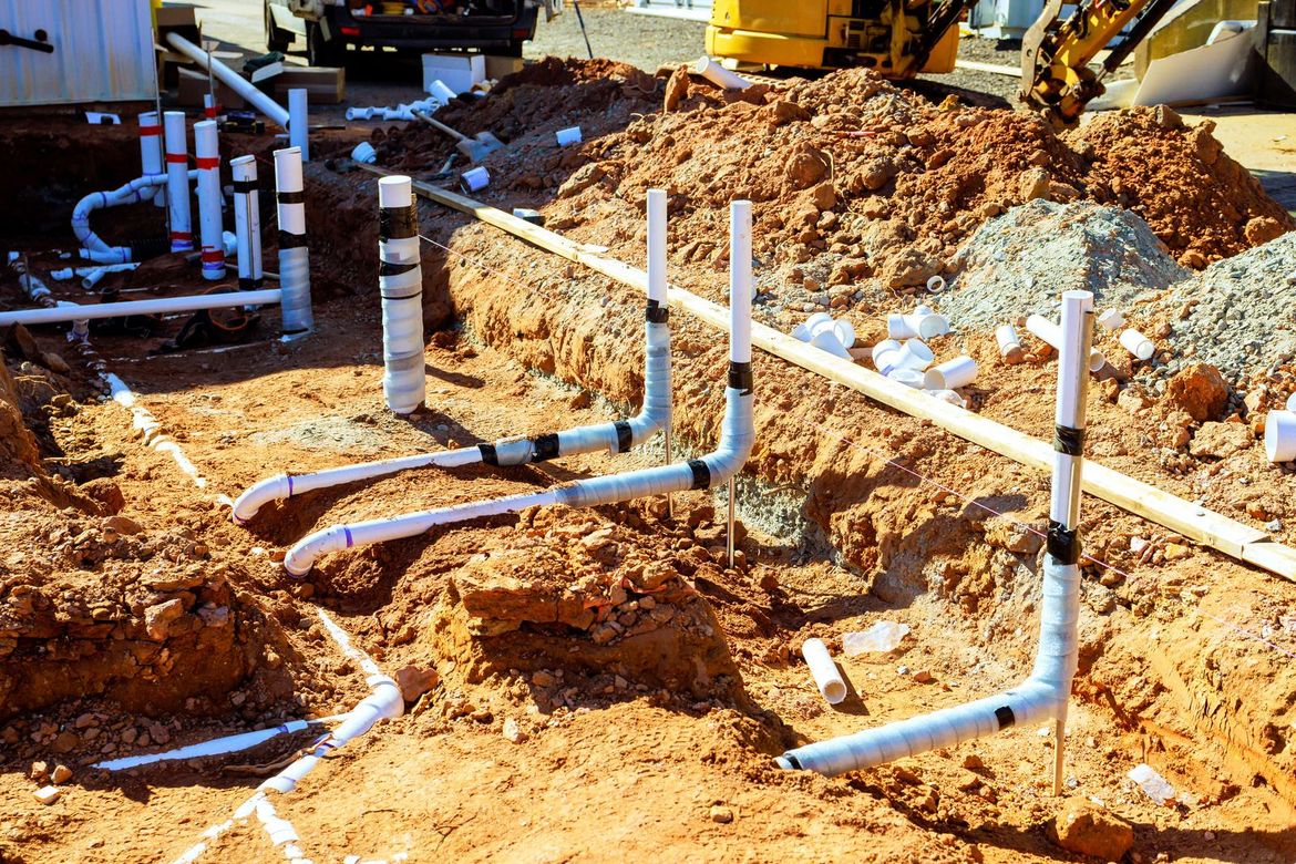 White PVC plumbing pipes emerge from a red dirt construction site with trenches and a yellow machine in the background.