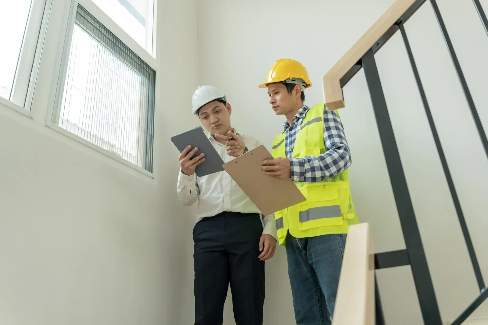 Two professionals in hard hats and work gear discuss construction plans using a tablet and clipboard in a building.