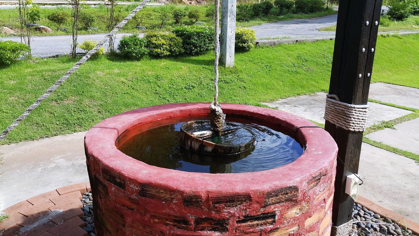 A red brick circular water well with a bucket hanging from a rope, situated on a lawn.