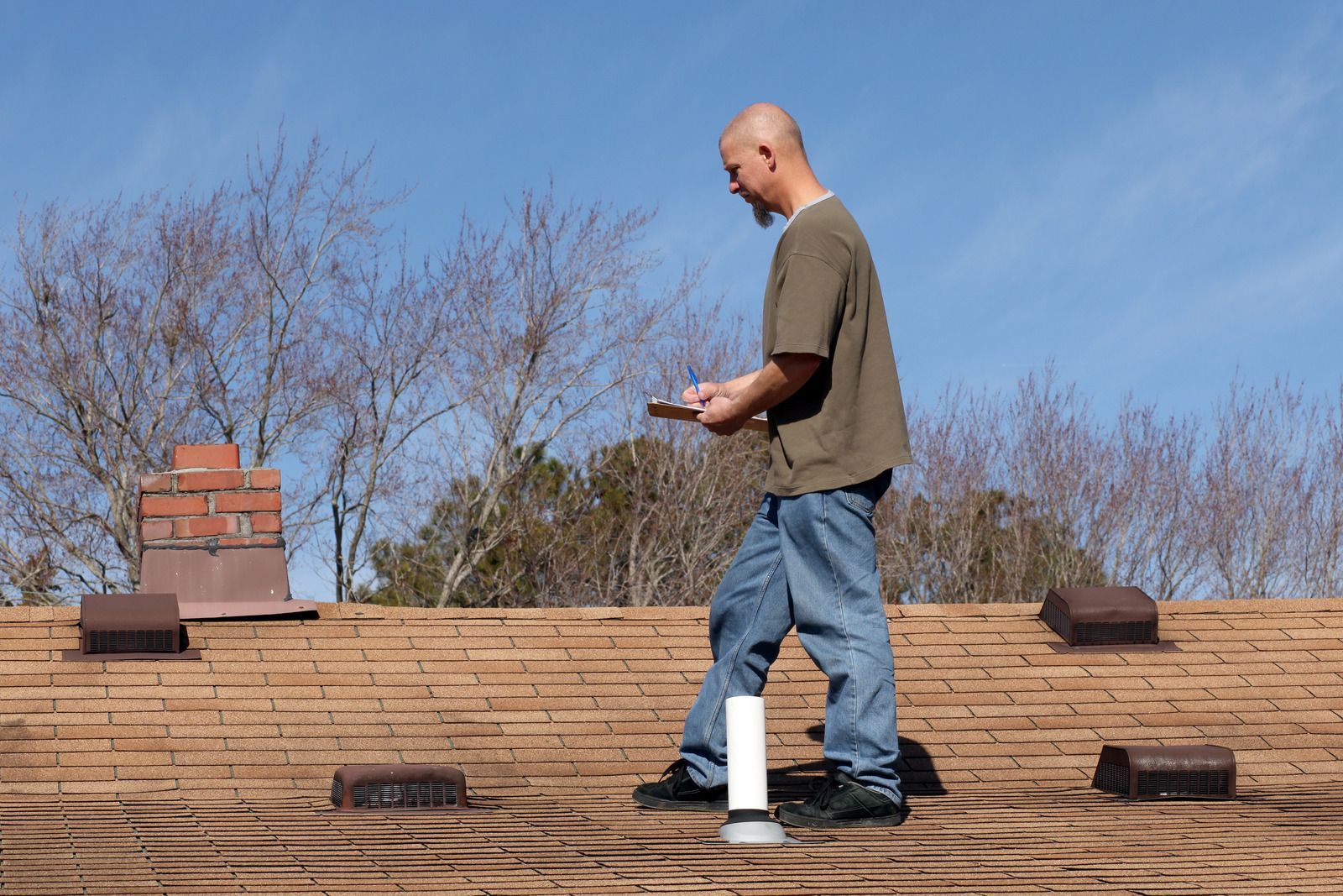 A person in a brown t-shirt and blue jeans walks across a brown shingled roof under a clear blue sky.
