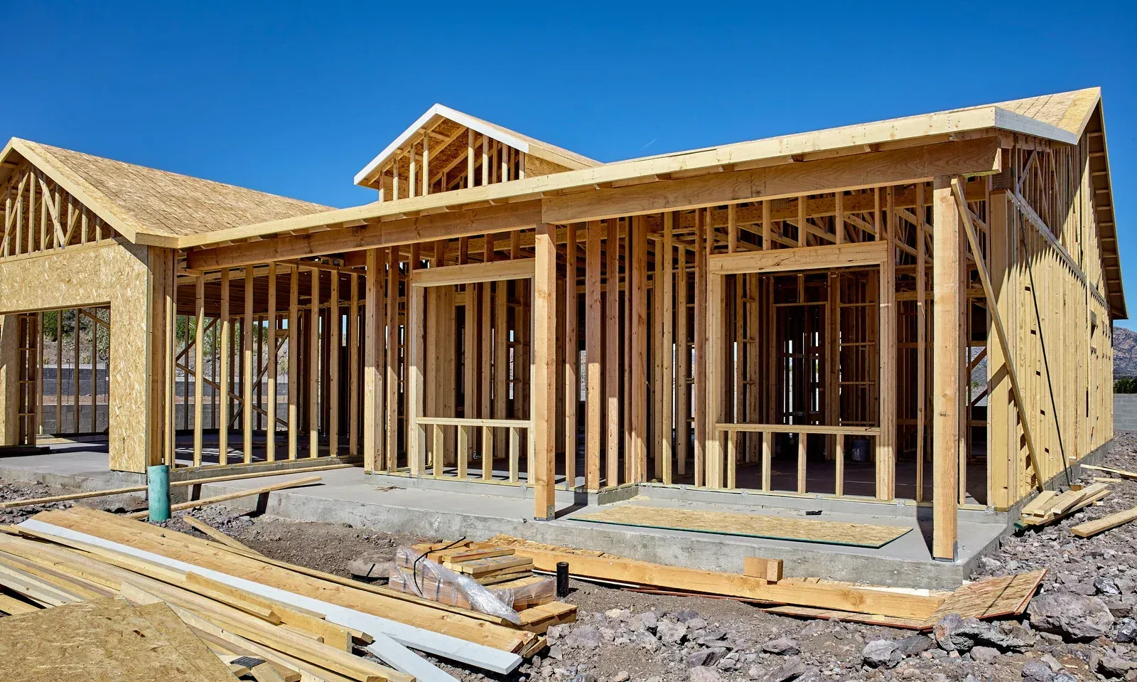 A wooden frame of a house under construction against a clear blue sky, with a pile of lumber in the foreground.