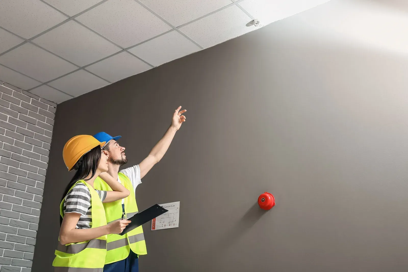 Two construction workers in safety vests and hard hats look up and point at a ceiling while holding a clipboard.
