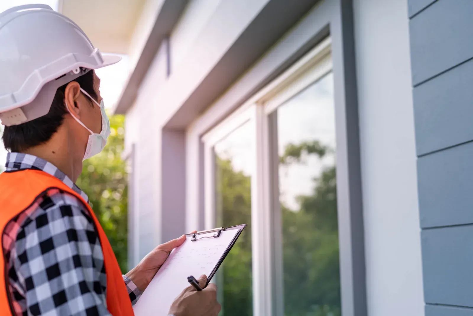 A construction worker in a hard hat and safety vest writing on a clipboard while inspecting a house window.