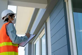 A person wearing a white hard hat and orange safety vest writes on a clipboard while inspecting a modern building exterior.