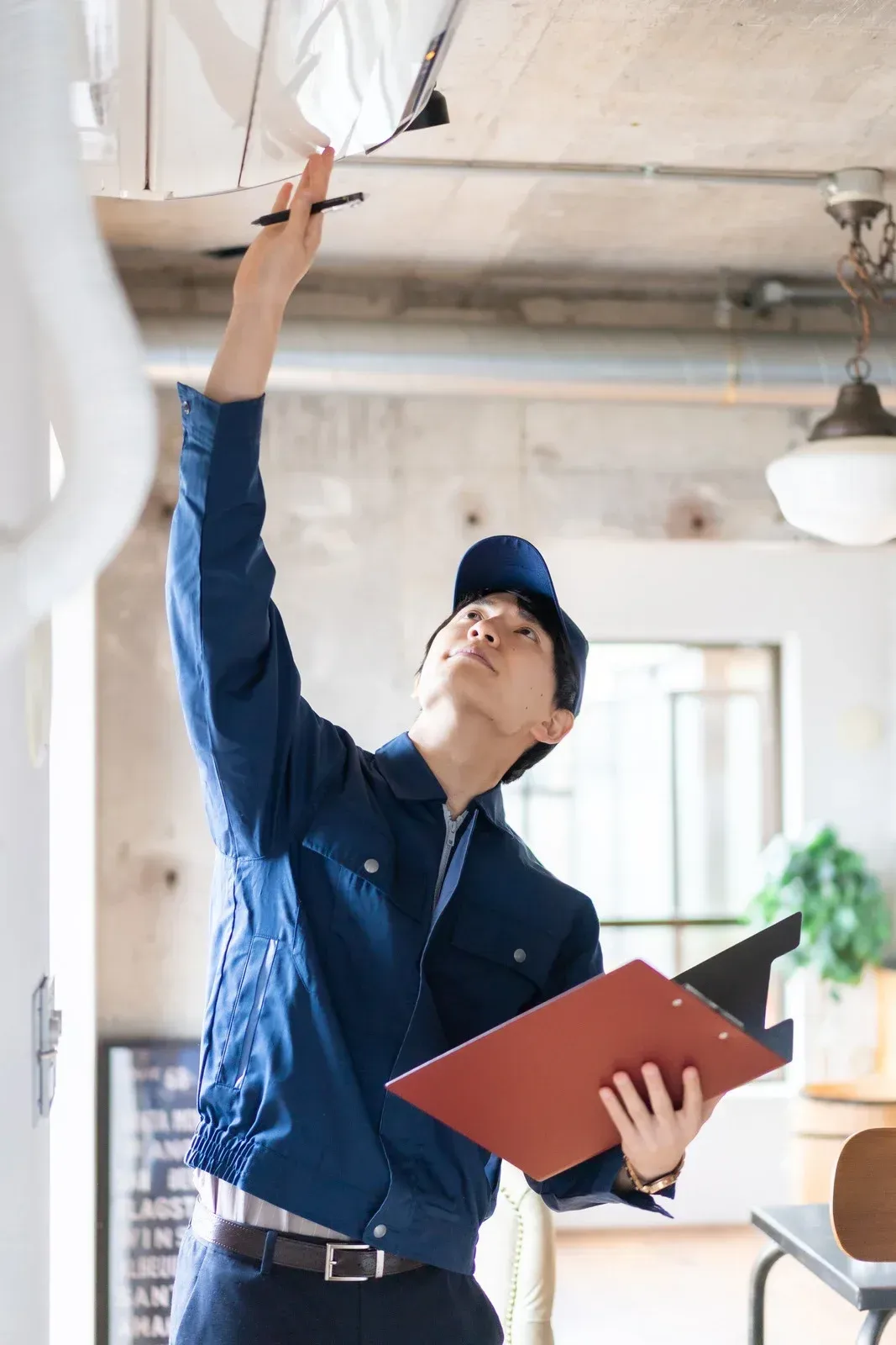 A person in a blue uniform and cap reaches up to inspect a ceiling fixture while holding a clipboard in an office.