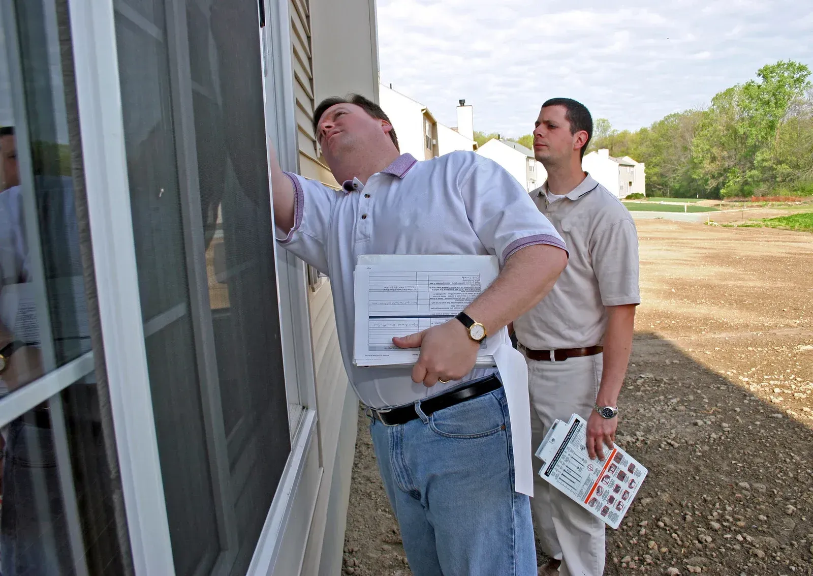 Two inspectors in casual clothing examine a sliding glass door at a construction site with clipboards in hand.