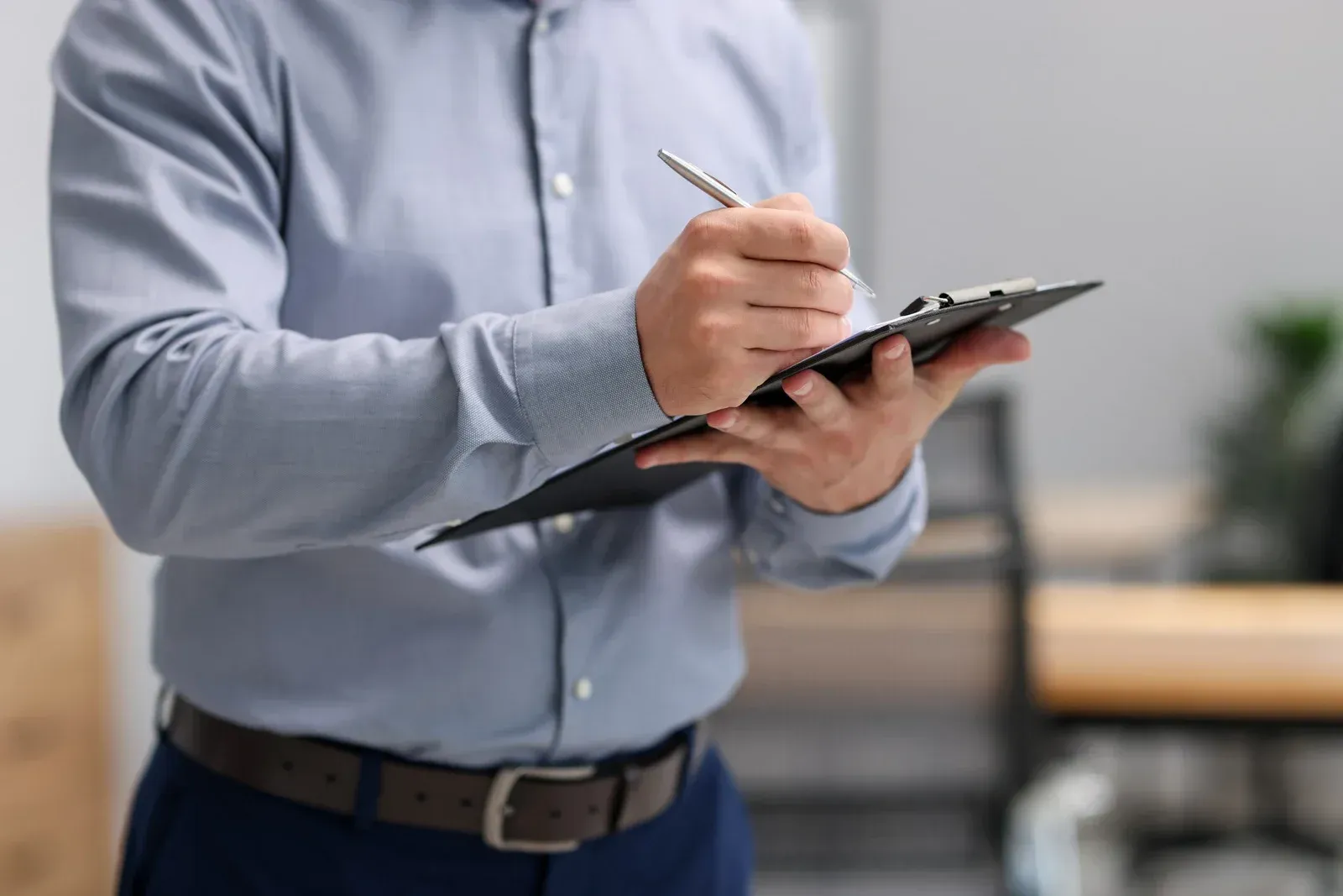 A person in a light blue button-down shirt and brown belt writes on a clipboard held in their hands in an office setting.