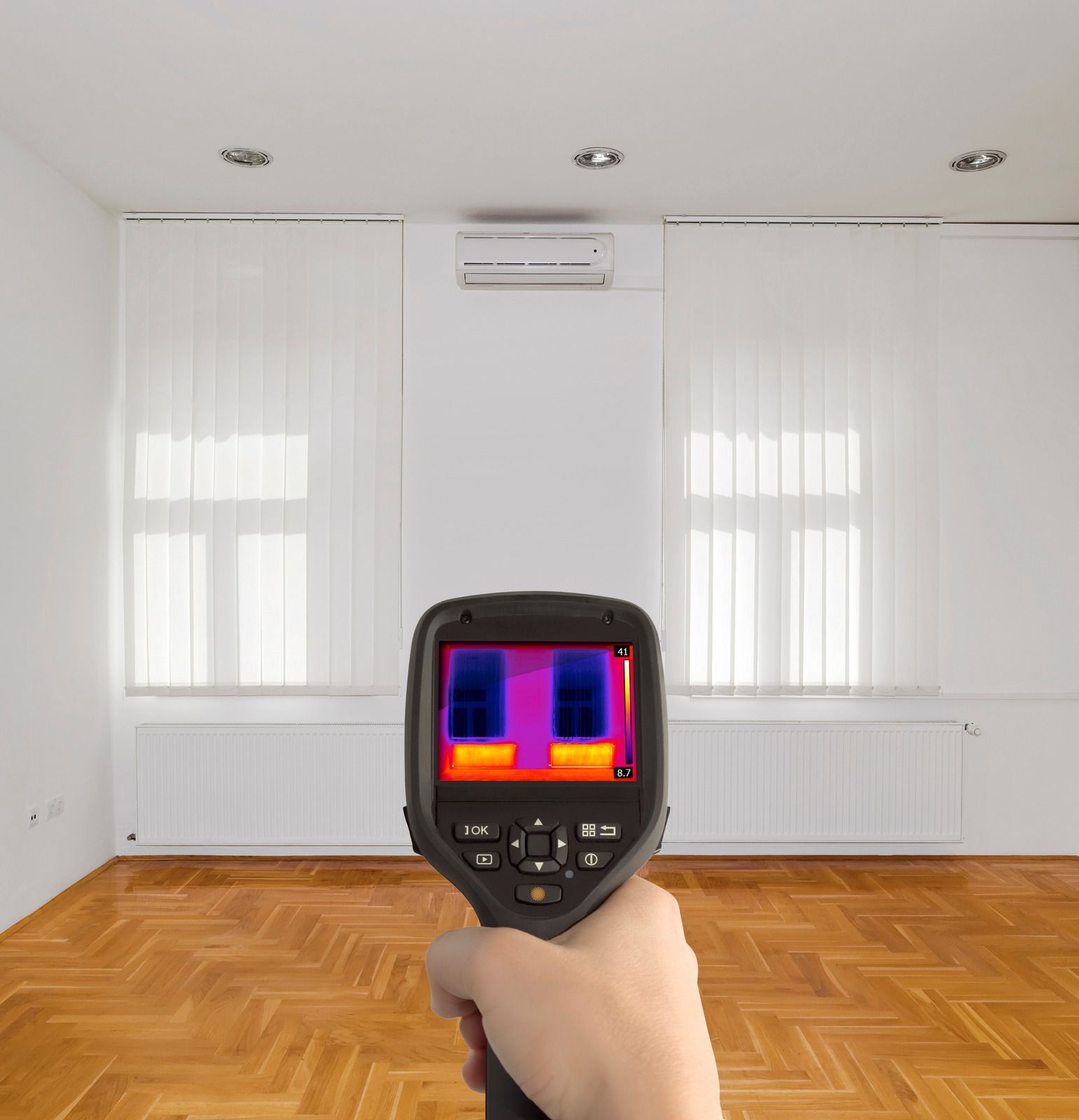 A person holds a thermal imaging camera in a room, displaying a heat map of windows in front of them.