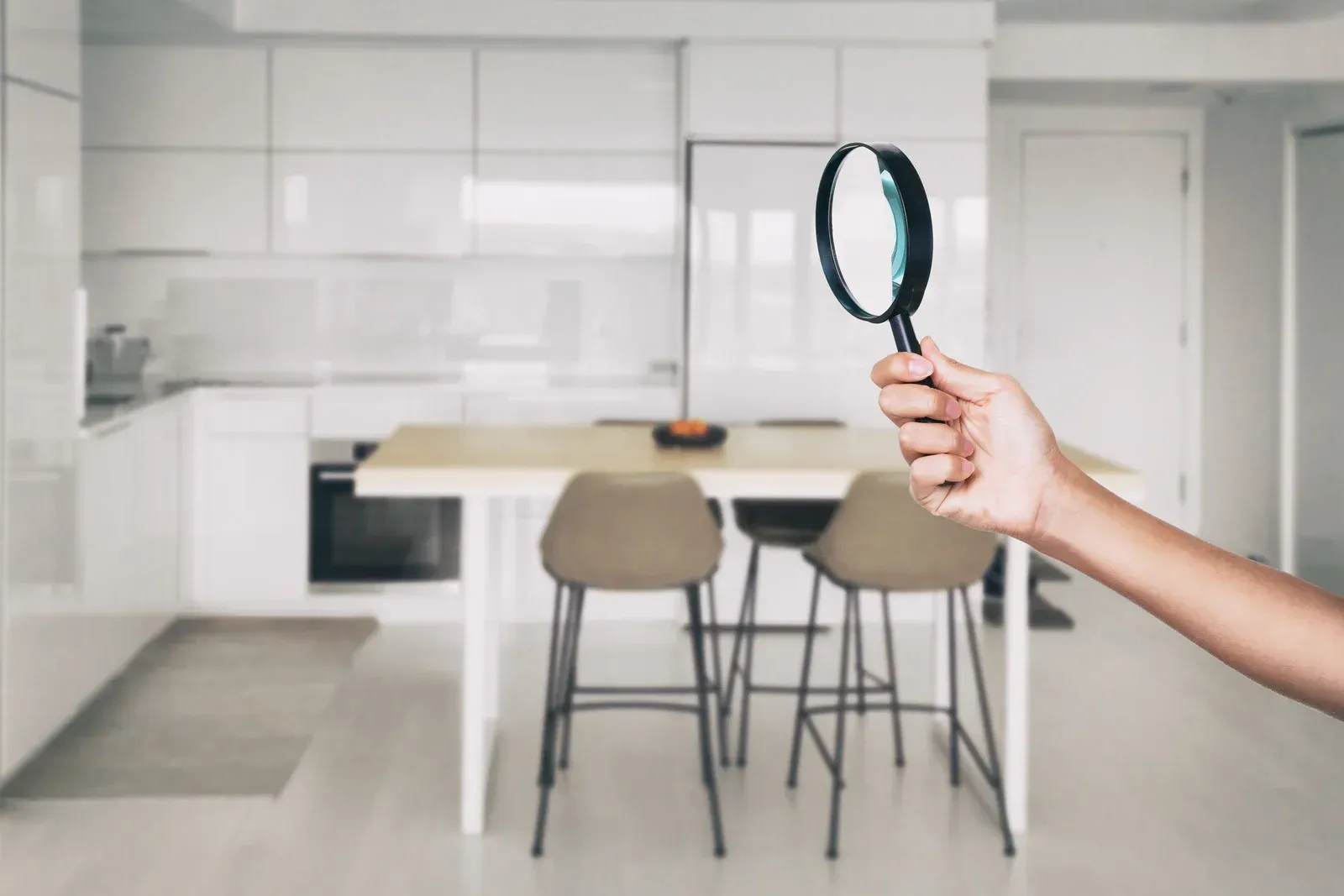 A hand holding a magnifying glass in front of a blurred, modern white kitchen interior.