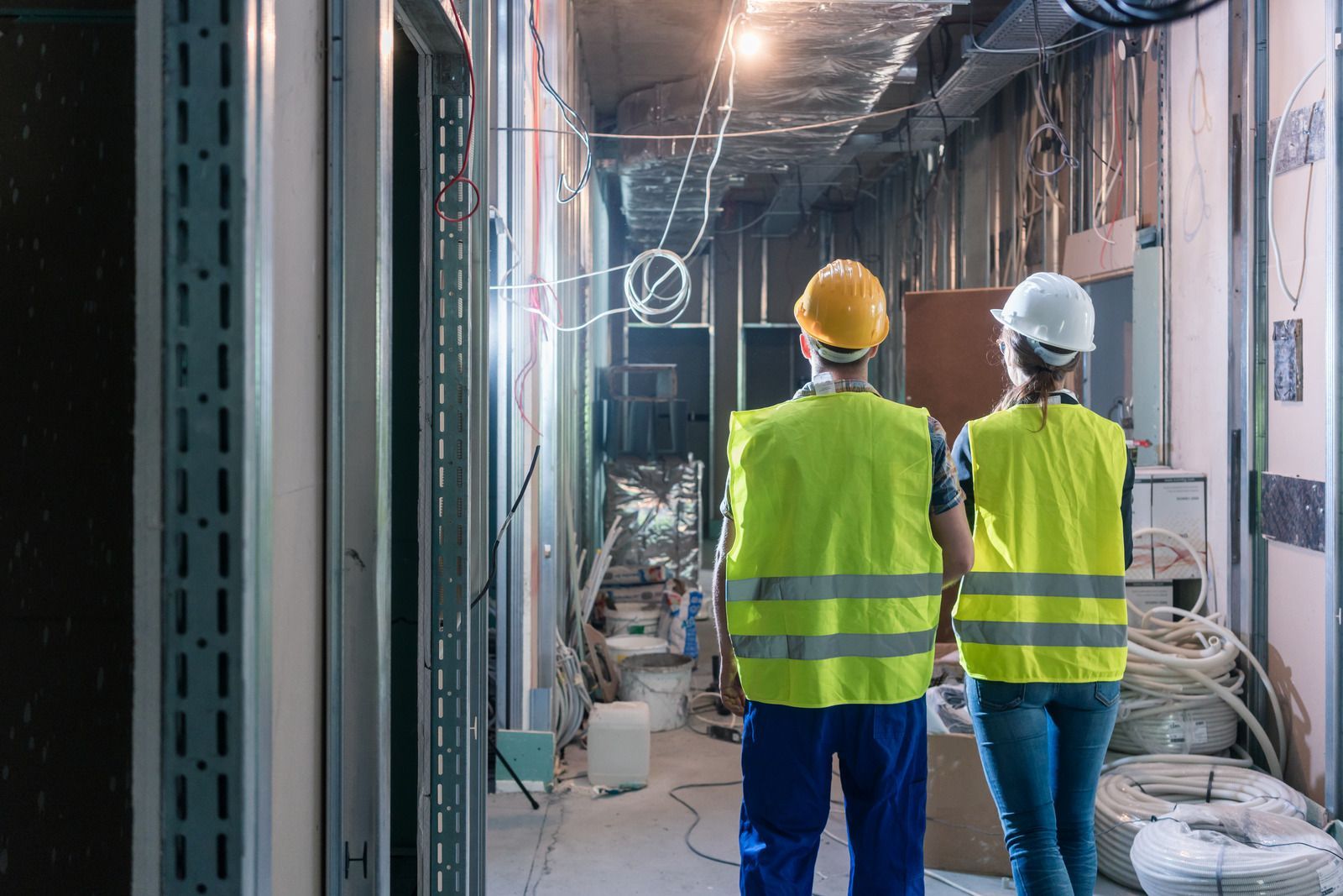 Two construction workers in high-visibility vests and hard hats stand in a hallway of an unfinished building.