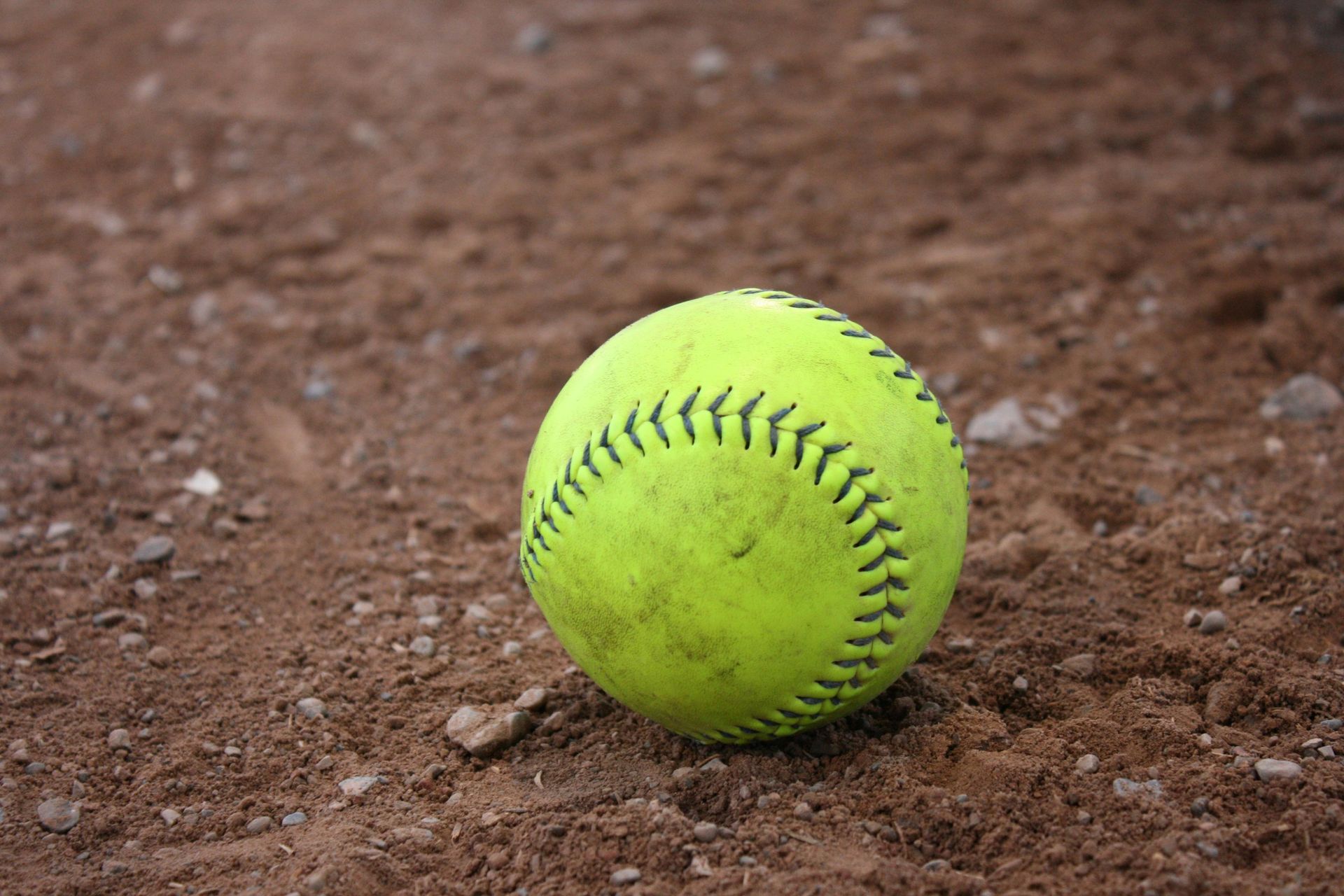 A bright yellow softball resting on a dirt field.