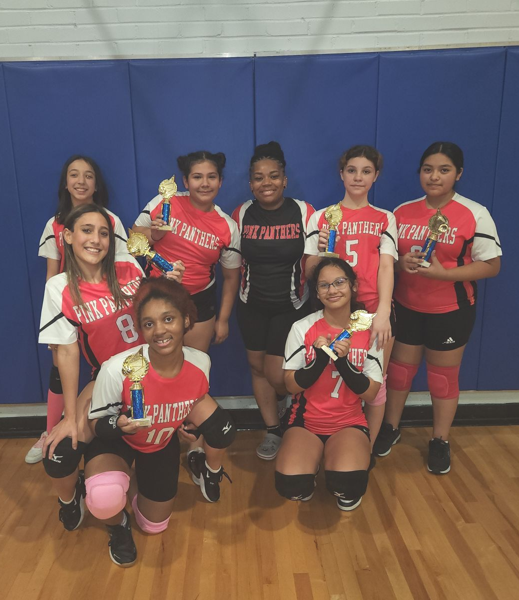 A group of eight volleyball players in red and white uniforms posing with trophies on a wooden court in front of a blue wall.