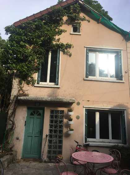 Maison à deux étages aux murs rosés, porte verte et plantes grimpantes. Table et chaises roses devant.
