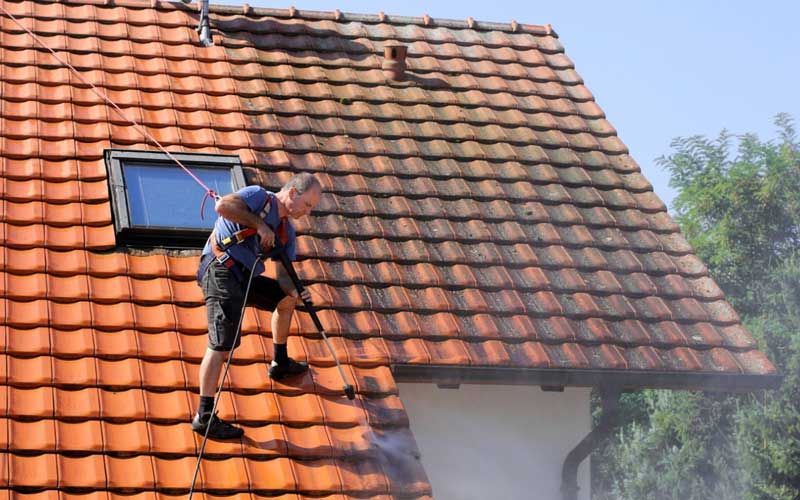 Une personne nettoie à haute pression un toit de tuiles rouges, sécurisée par un harnais et une corde.