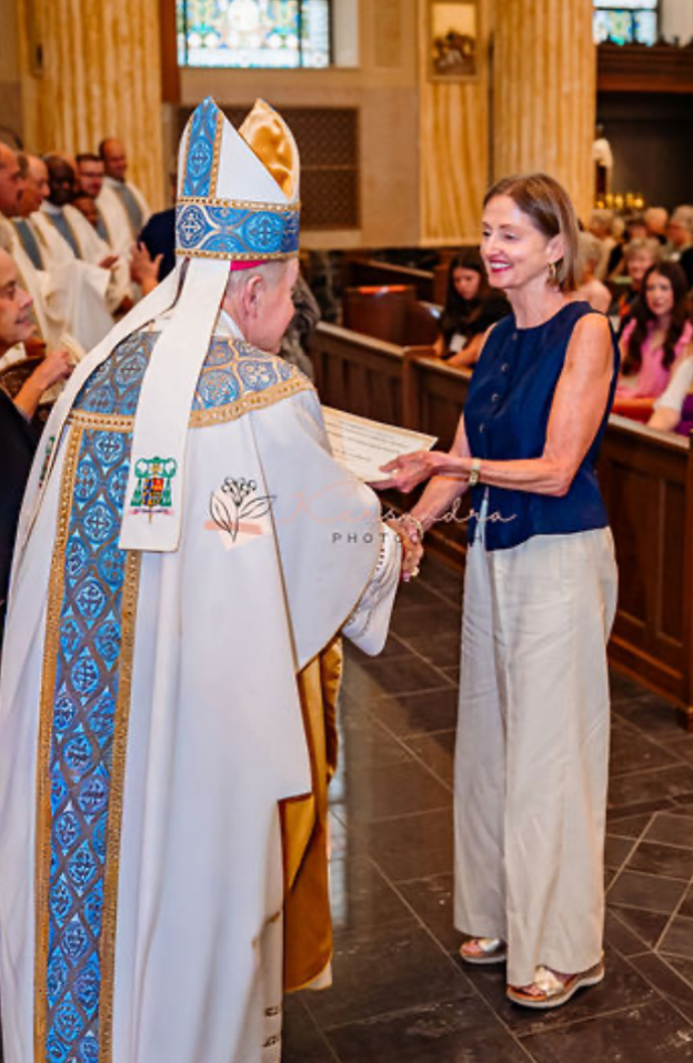 Bishop in white vestments hands a booklet to a smiling woman in a church aisle