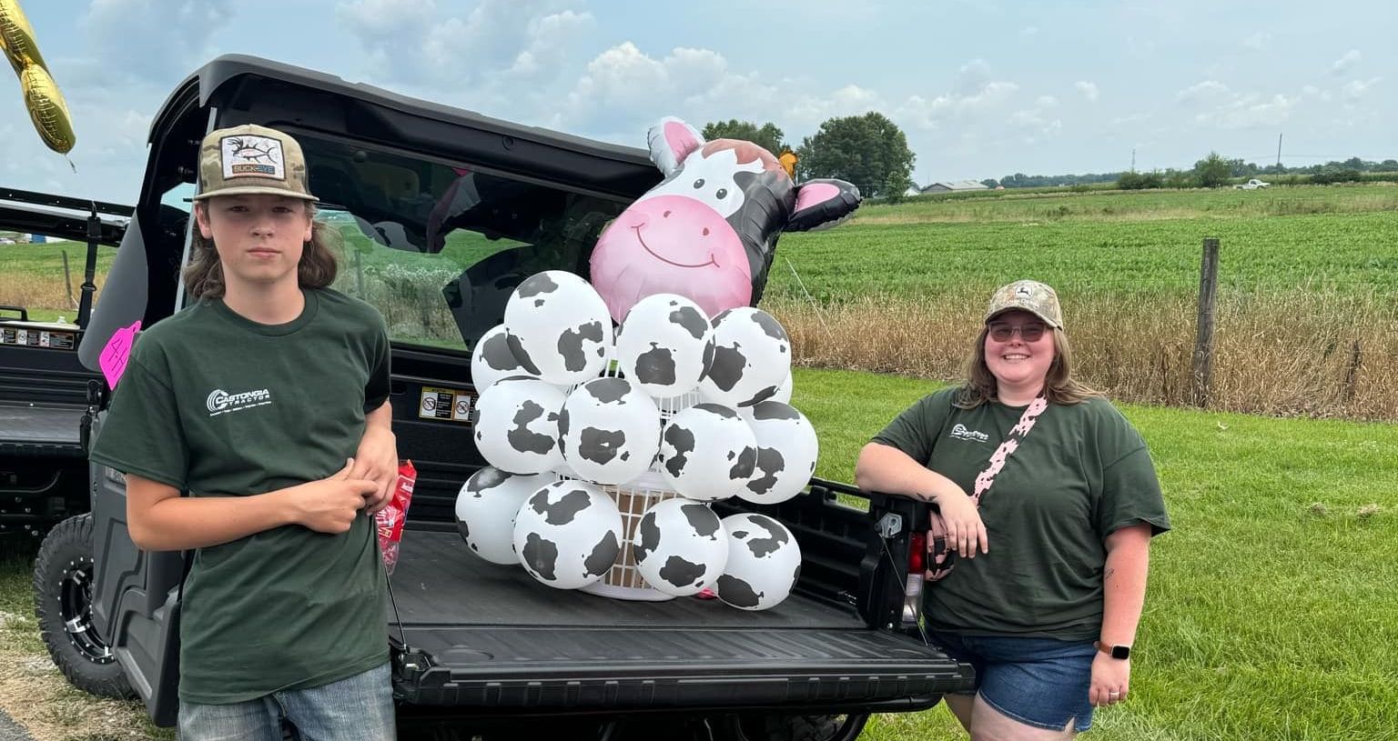 one male, one female pose by the tailgate of a John Deere Gator. the bed of the UTV has a balloon arrangement of a dairy cow.