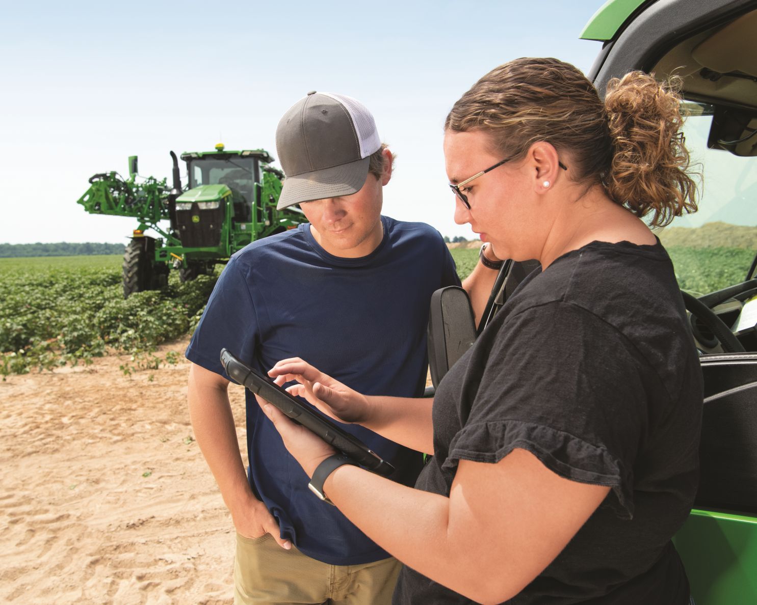 man and woman look at an iPad with a John Deere sprayer parked behind them in a field