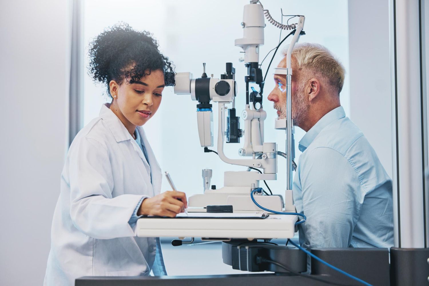 Optometrist examining a man's eye with a slit lamp, writing notes in an office.