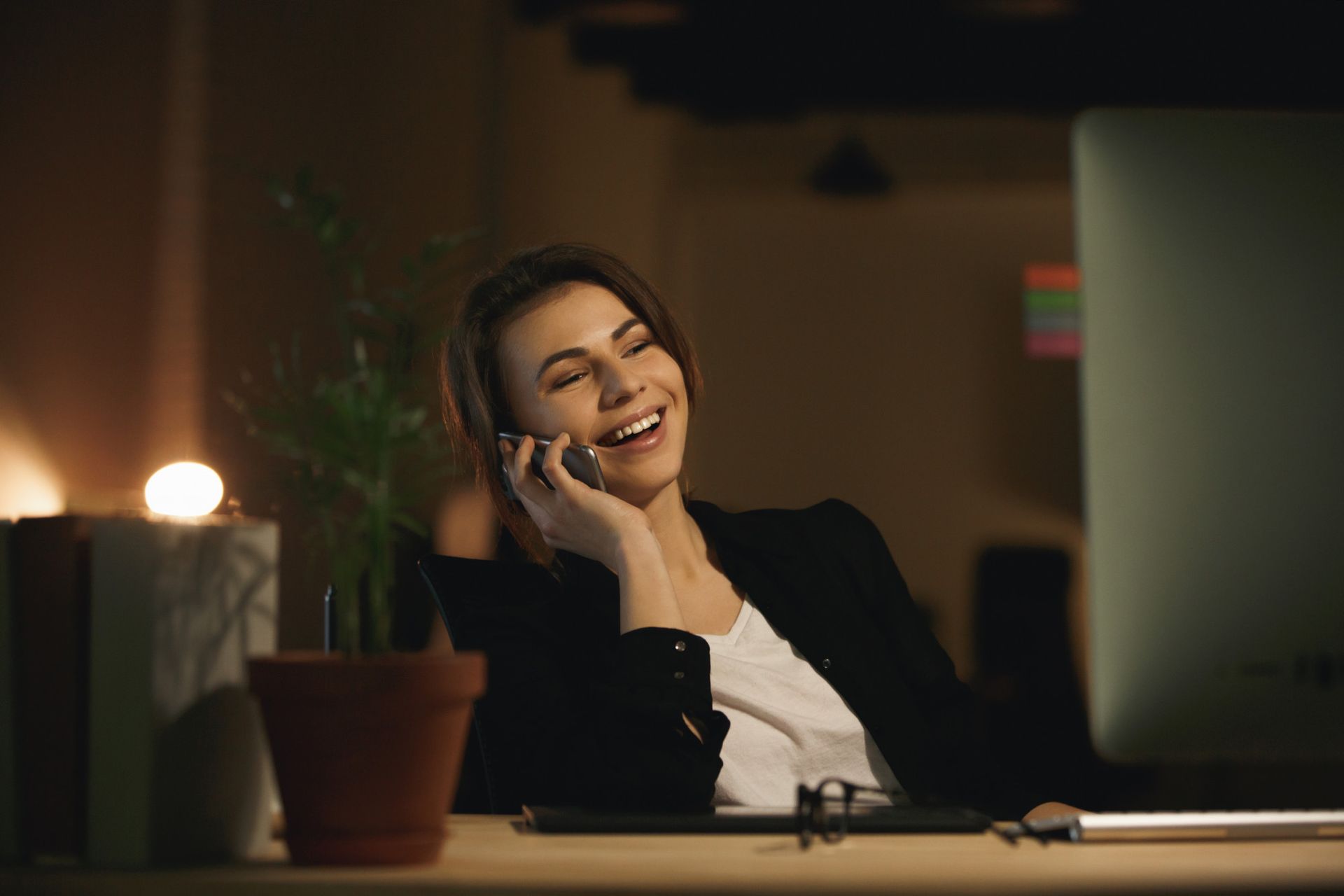 Woman in a dark office, laughing while on a phone call.