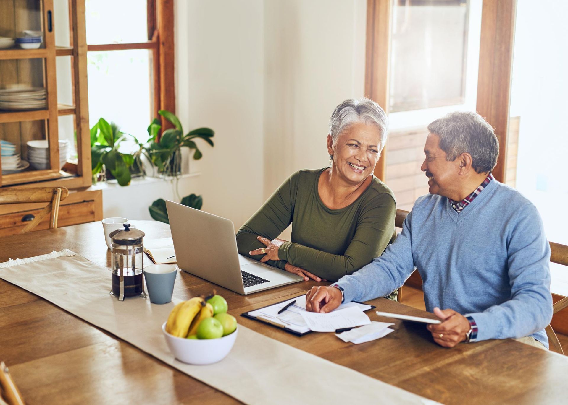Older couple reviewing finances together at a wooden table with a laptop, papers, and a tablet.