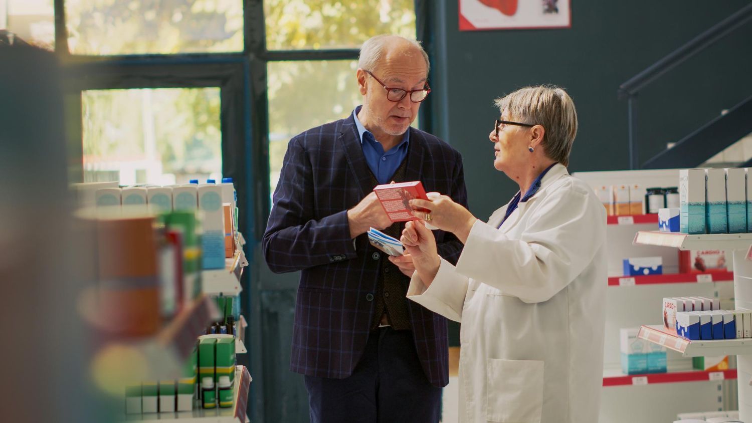Pharmacist in white coat helps elderly man at pharmacy counter, showing him medicine box.