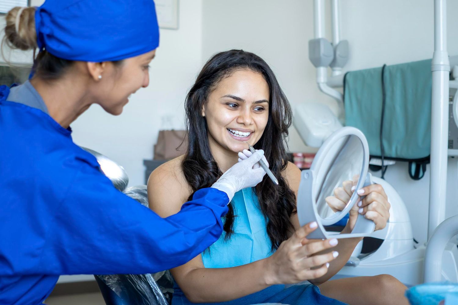Dentist in blue scrubs showing a patient her teeth in a mirror at a dental office.
