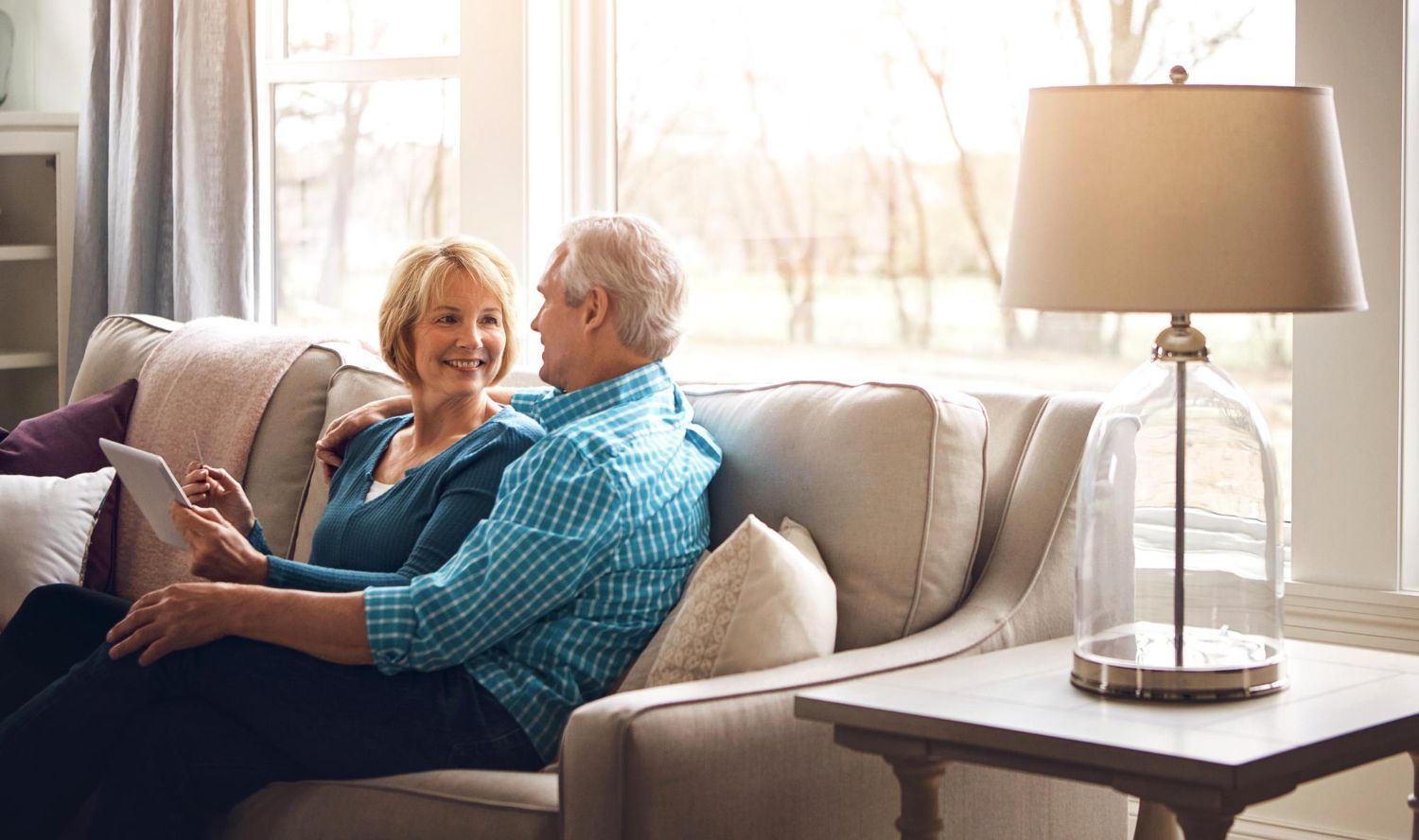An older couple smiles while sitting on a sofa, looking at a tablet in a living room.