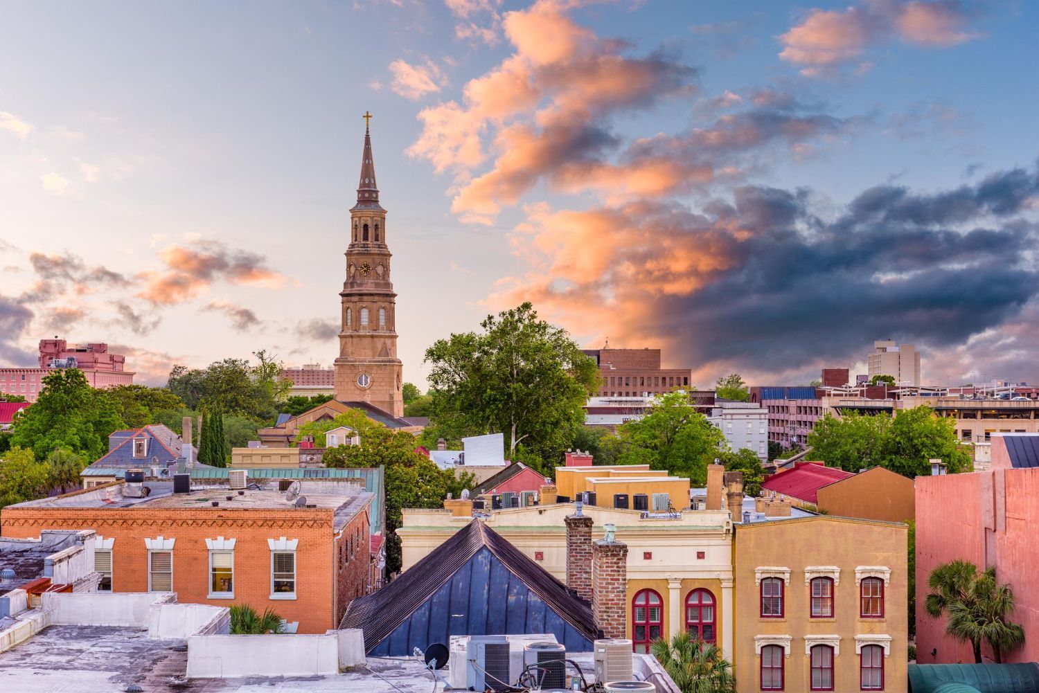 Rooftop view of Charleston, SC, with colorful buildings, a church steeple, and a sunset sky.