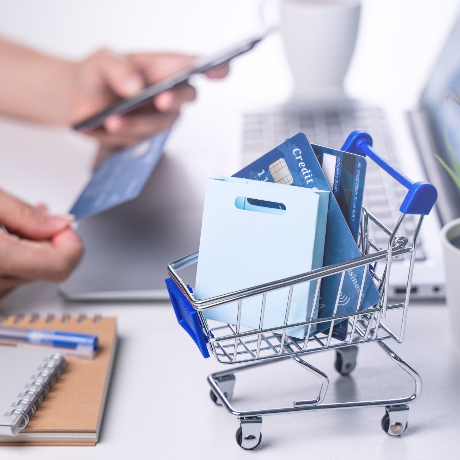 Shopping cart with credit cards and gift bag near a person shopping on a phone and laptop.