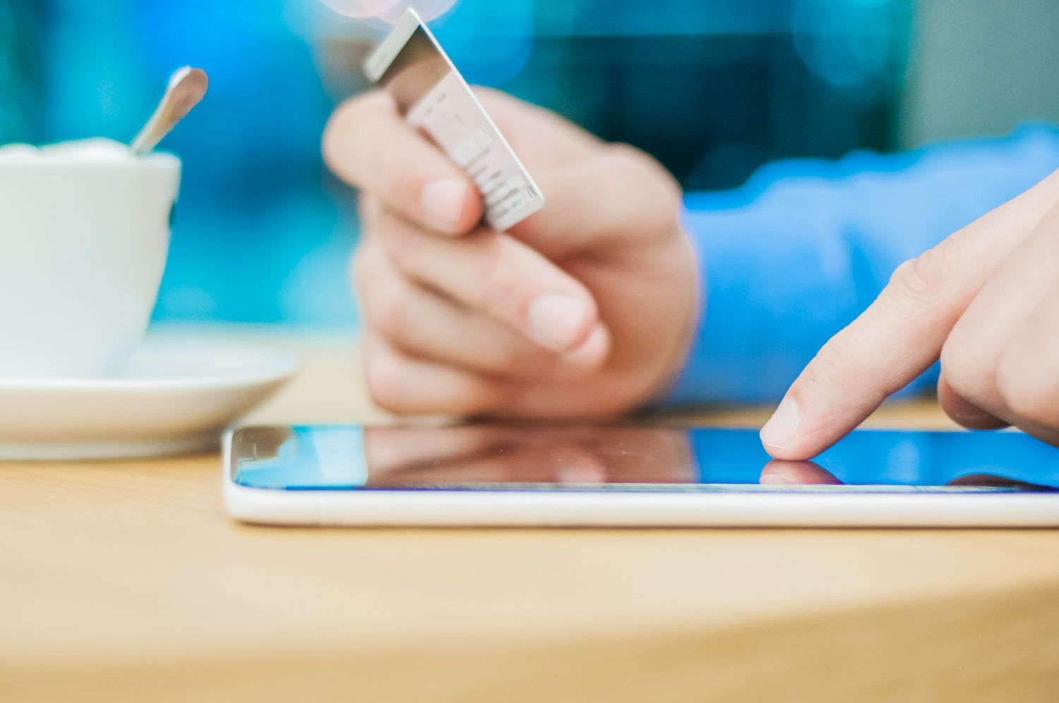 Person uses a tablet and credit card at a table, likely for online shopping. Close-up view with coffee cup present.