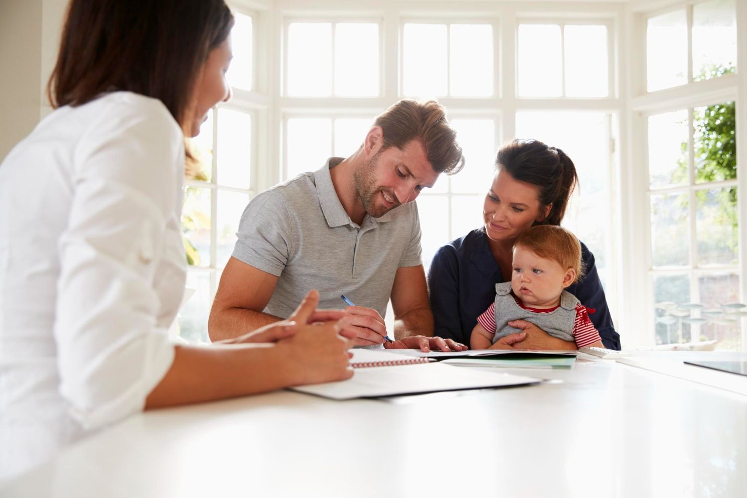 Woman advising a family: man signing documents, woman and baby looking on, at table, near window.