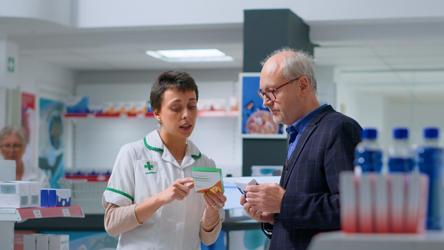 Pharmacist in white coat explains medication to a man in a pharmacy setting.