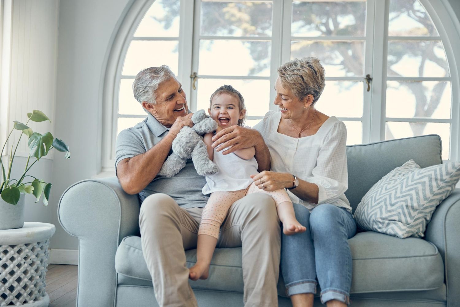 Grandparents and a child laughing on a couch. Grandfather holds a toy; grandmother smiles in a bright living room.