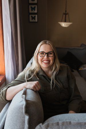 A woman wearing glasses is sitting on a couch and smiling.