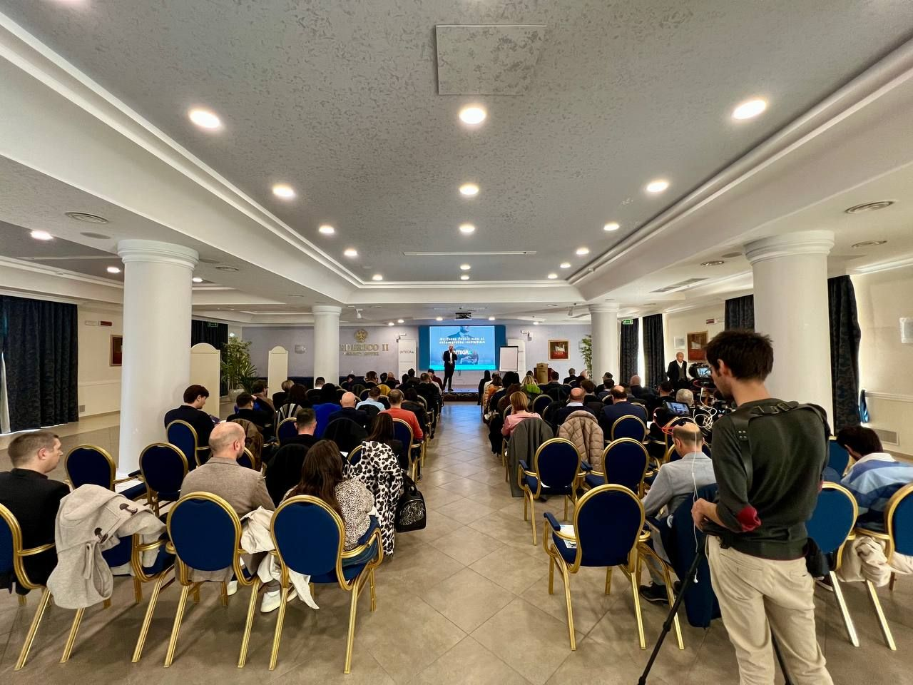 Una sala conferenze con un relatore in prima fila, file di partecipanti e un uomo che filma.