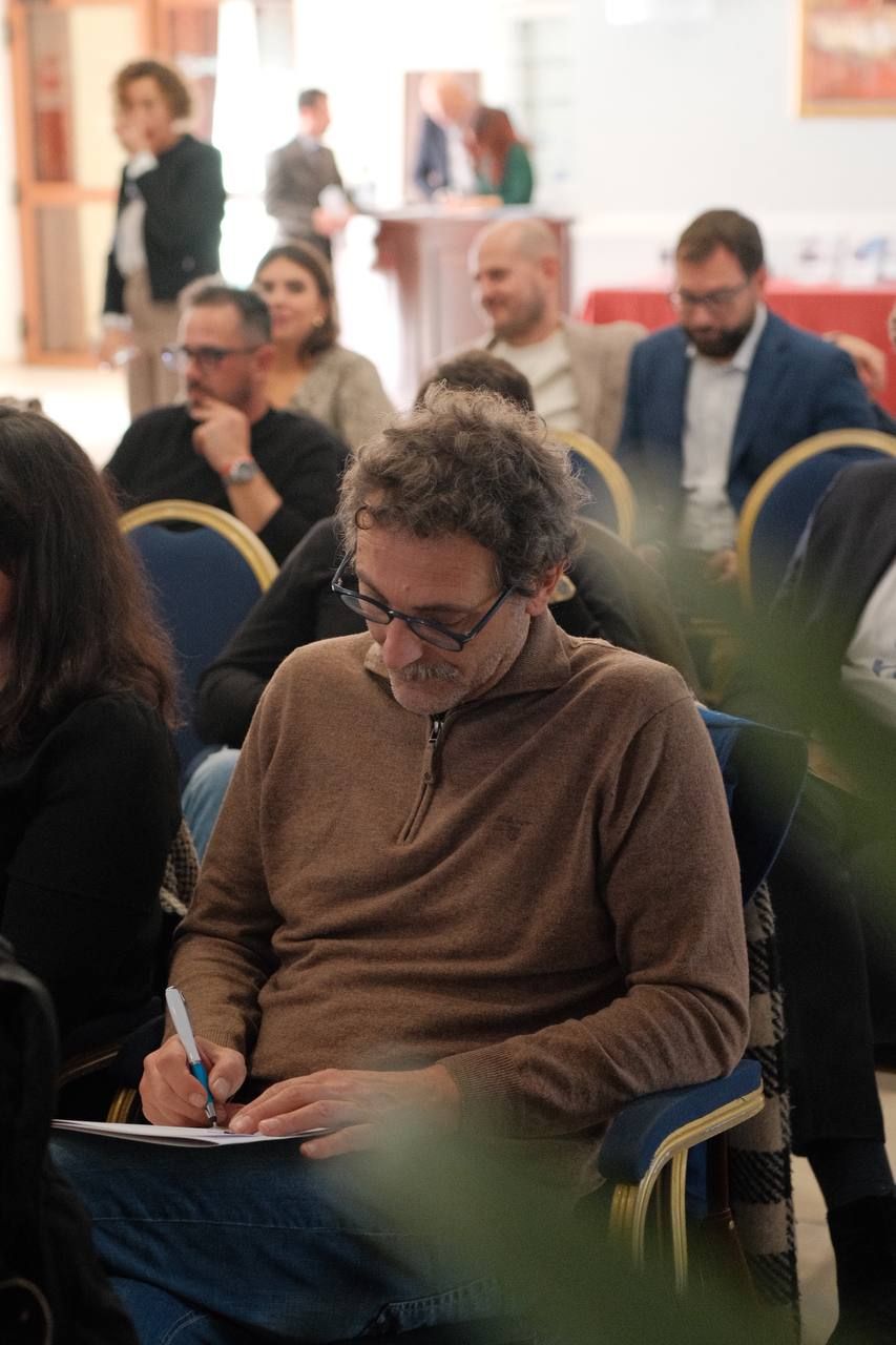 Un uomo con gli occhiali scrive su un blocco note durante un evento, circondato dagli altri partecipanti.