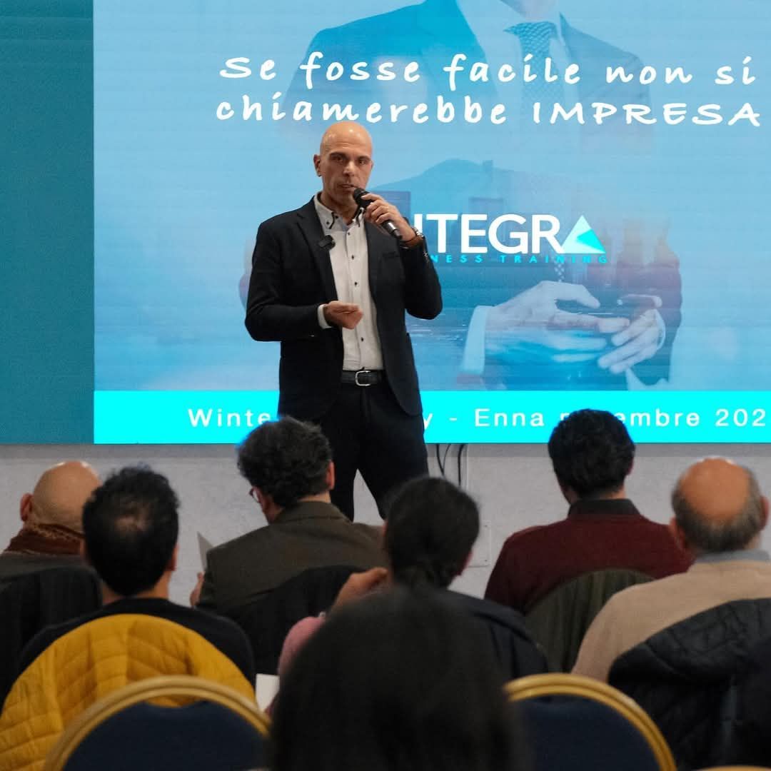 Uomo che parla a una conferenza. Il pubblico è seduto e guarda. Logo Integra sullo schermo, testo in italiano.