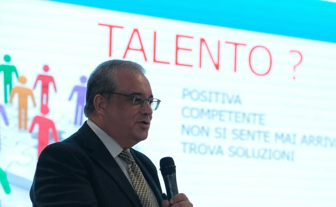 Un uomo parla al microfono mentre sullo schermo alle sue spalle compare il testo: 