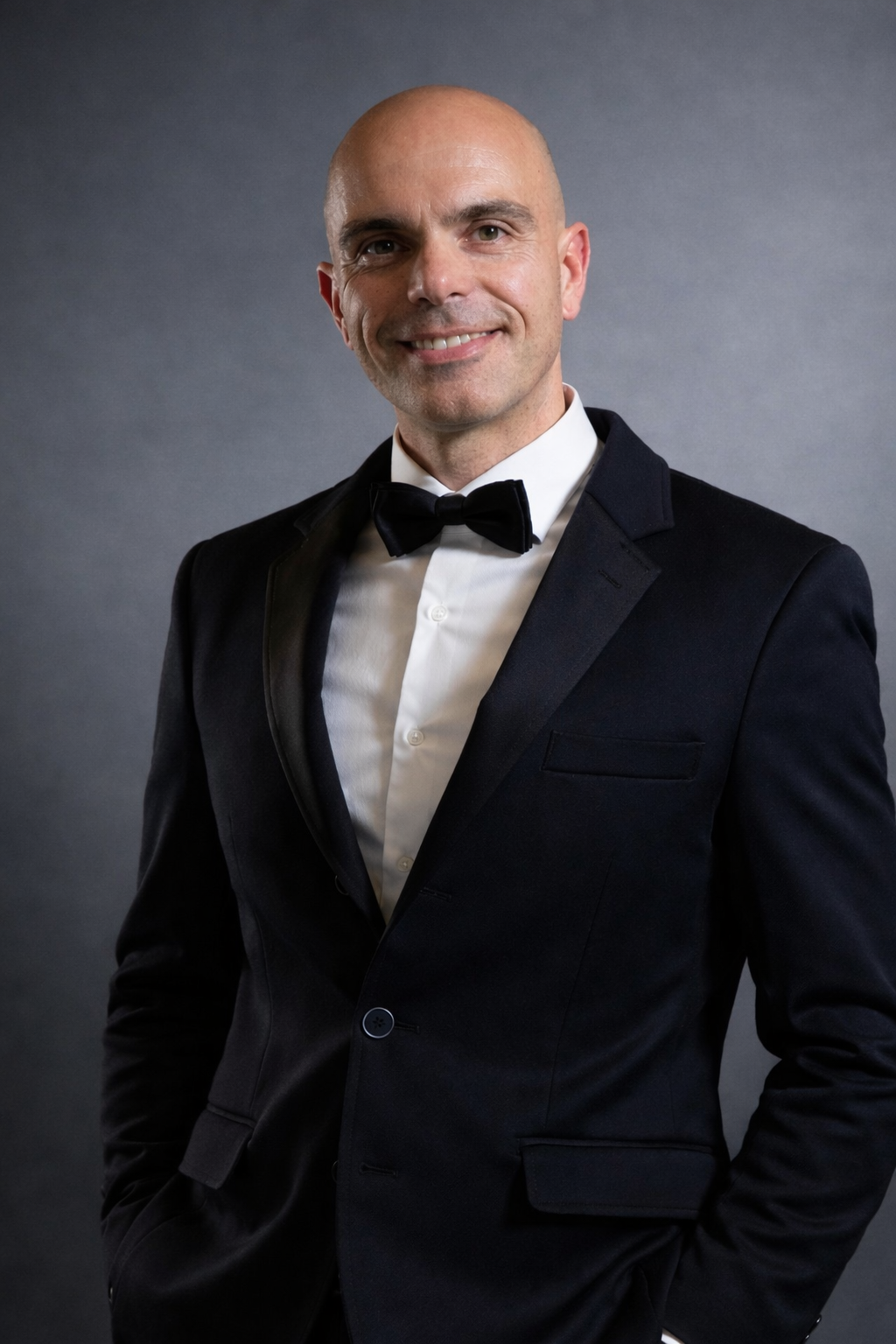 Uomo in smoking nero con papillon, sorridente, mani in tasca, sfondo grigio.