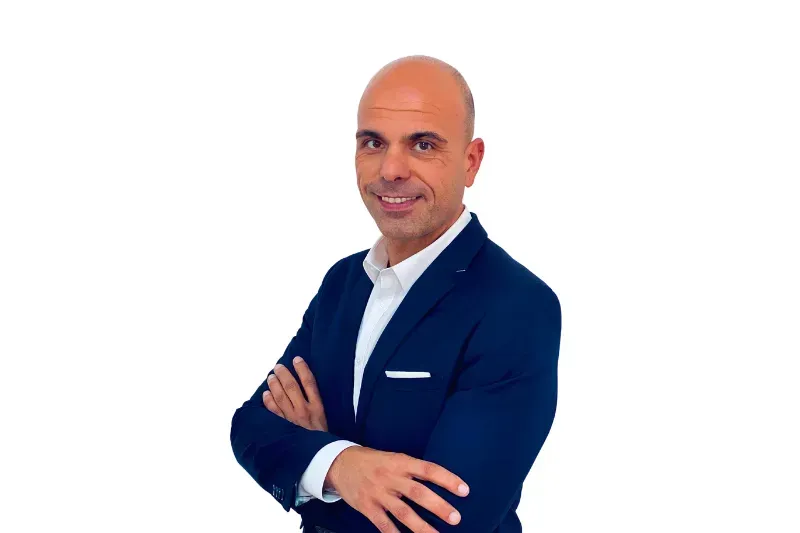 Uomo calvo con le braccia incrociate, vestito con un abito blu navy e una camicia bianca, sorridente.