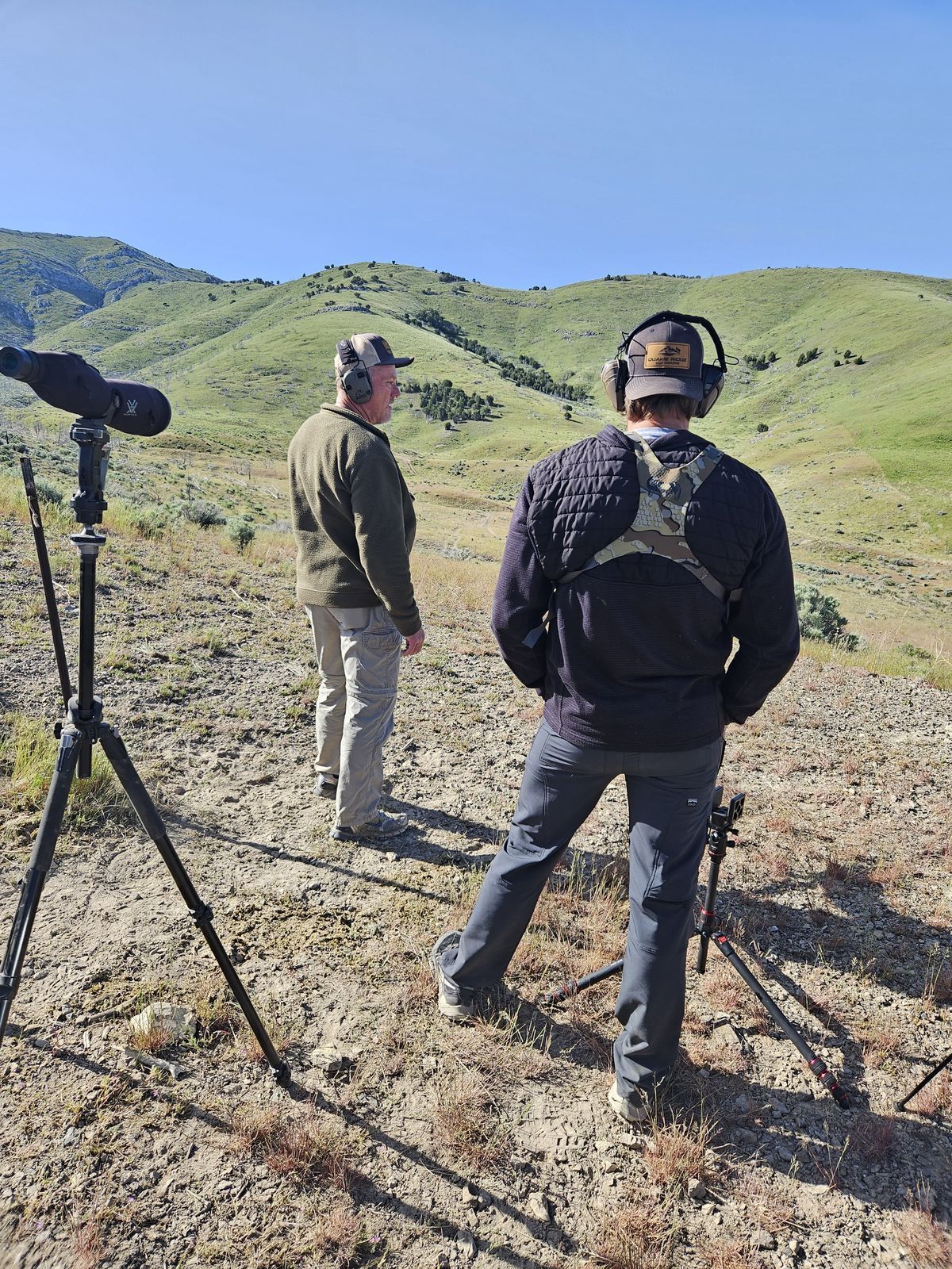 Two men wearing headphones are standing in a field with mountains in the background.