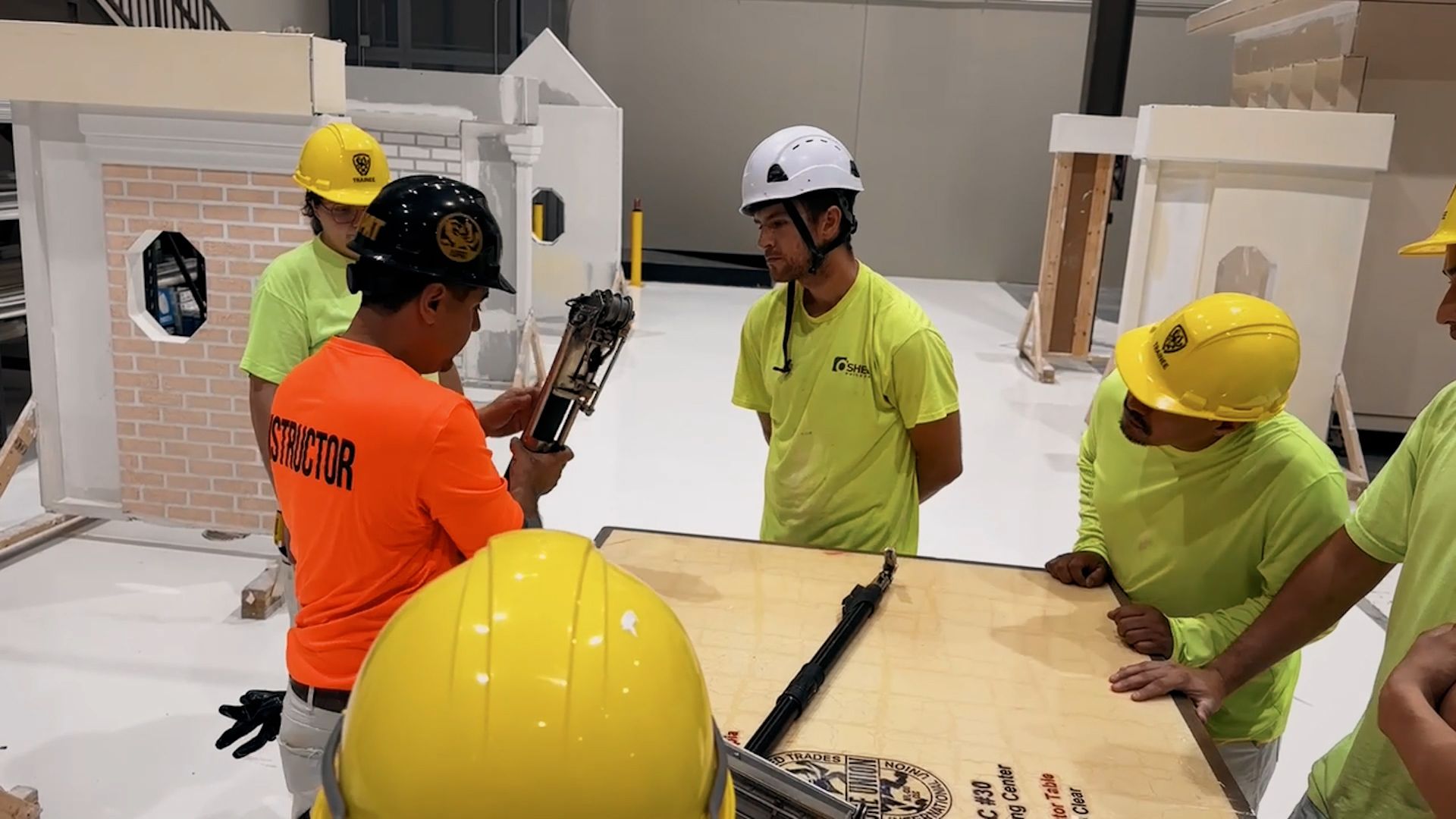 An NCIFTI instructor in an orange shirt demonstrating technical measurements to a group of students gathered around a table.