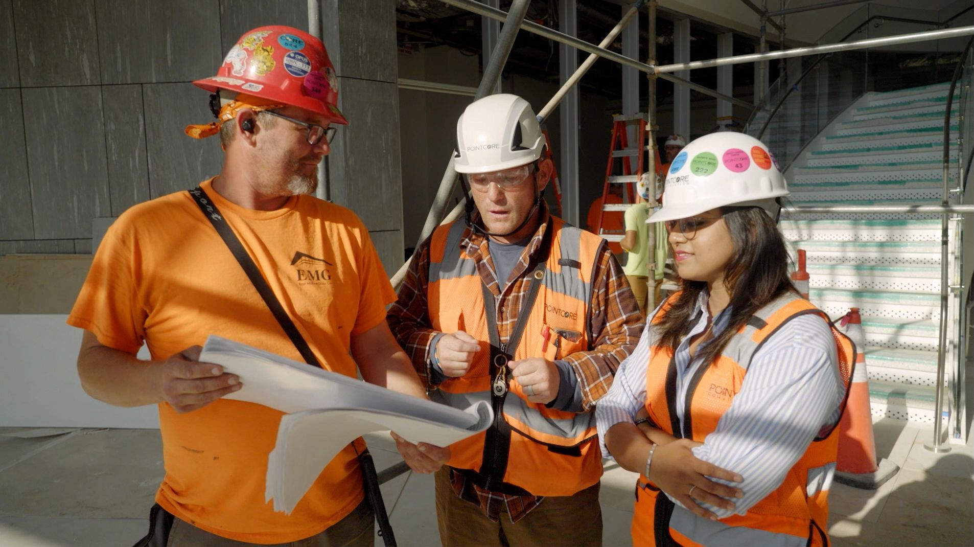 Three tradespeople on a job site wearing hard hats and safety vests, collaborating and reviewing architectural blueprints.