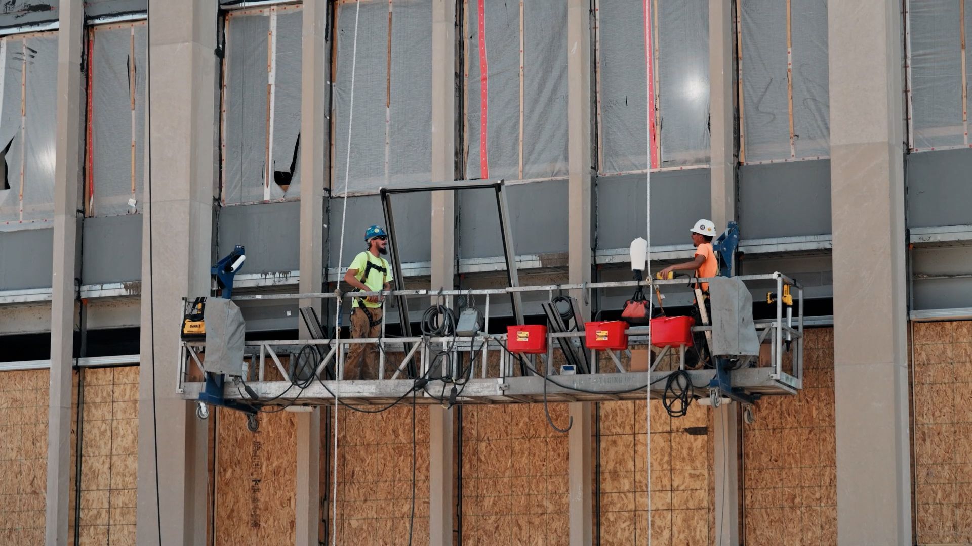 Two NCIFTI glazier workers from a suspended swing stage to install glass panels on a building's exterior.