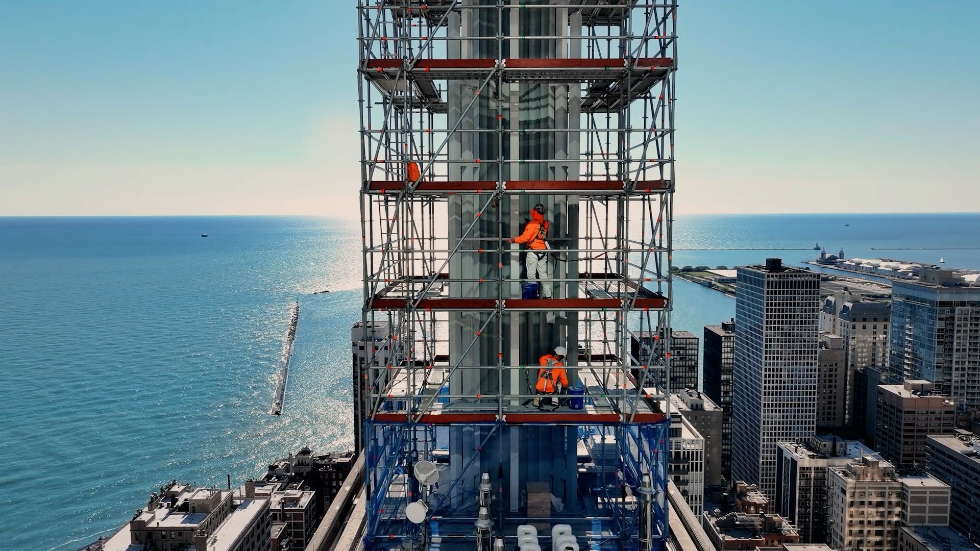 An aerial view of industrial painters working on a high-rise scaffolding structure overlooking the city coastline.