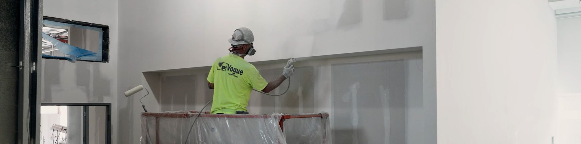 An NCIFTI apprentice in protective gear using an industrial sprayer to apply a smooth white finish to a large wall.