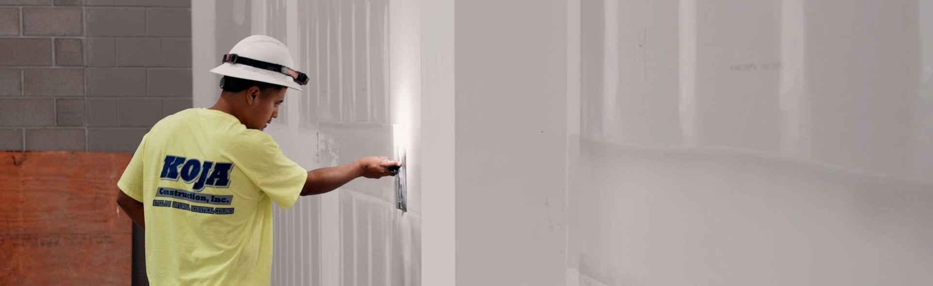 A close-up of an apprentice in a hard hat and headlamp using a finishing knife for detailed drywall work.