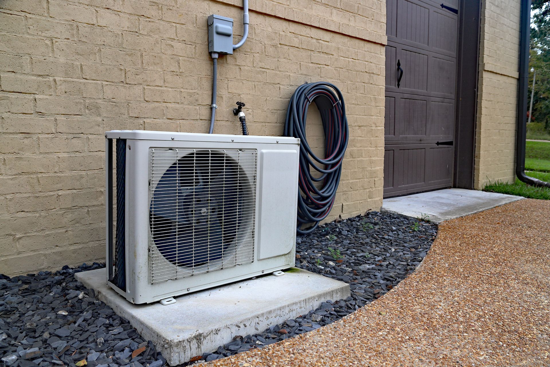 A white air conditioner is sitting outside of a brick building next to a hose.