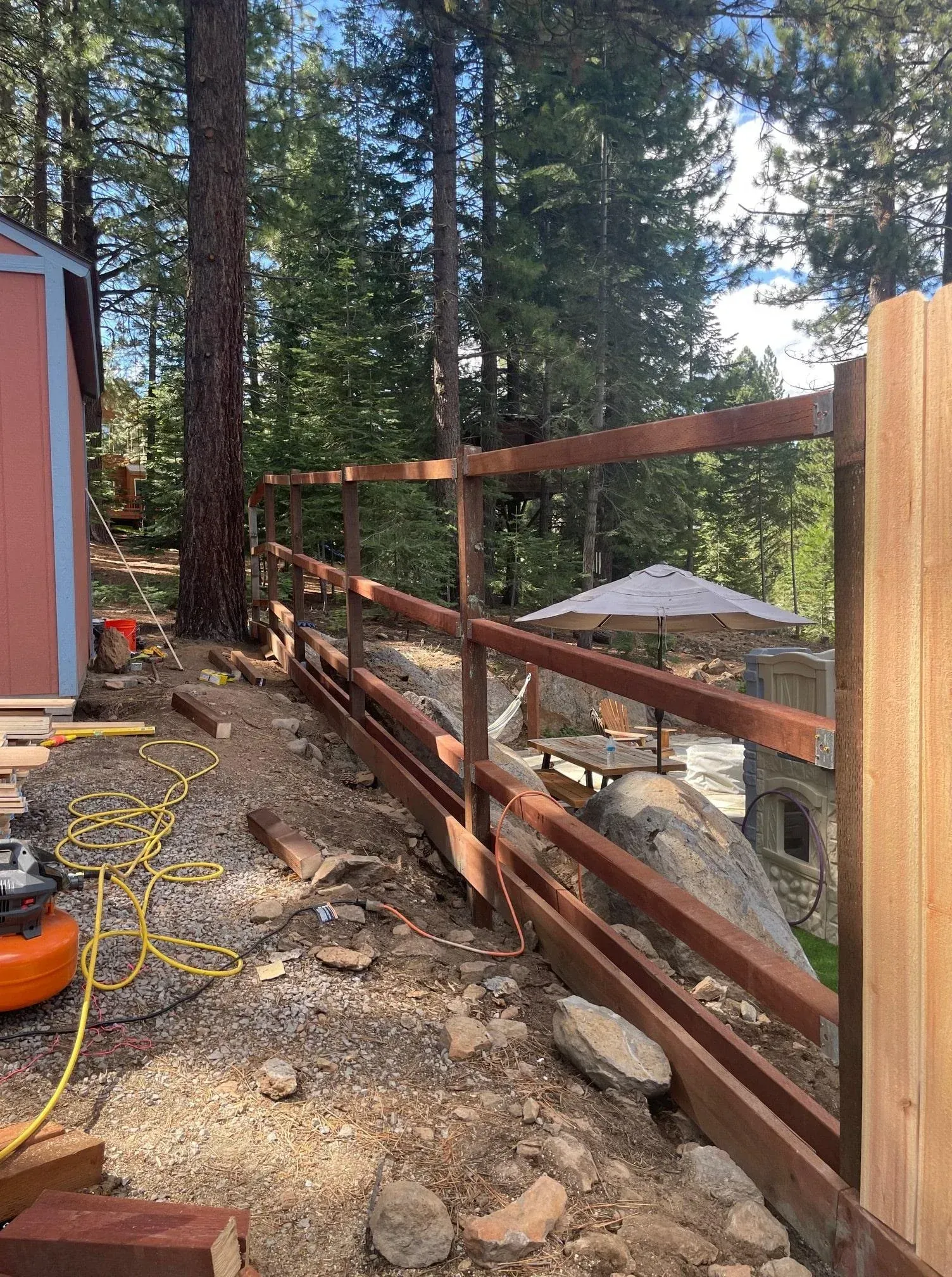Wooden fence being built on a hillside, overlooking a patio with a white umbrella, surrounded by trees.