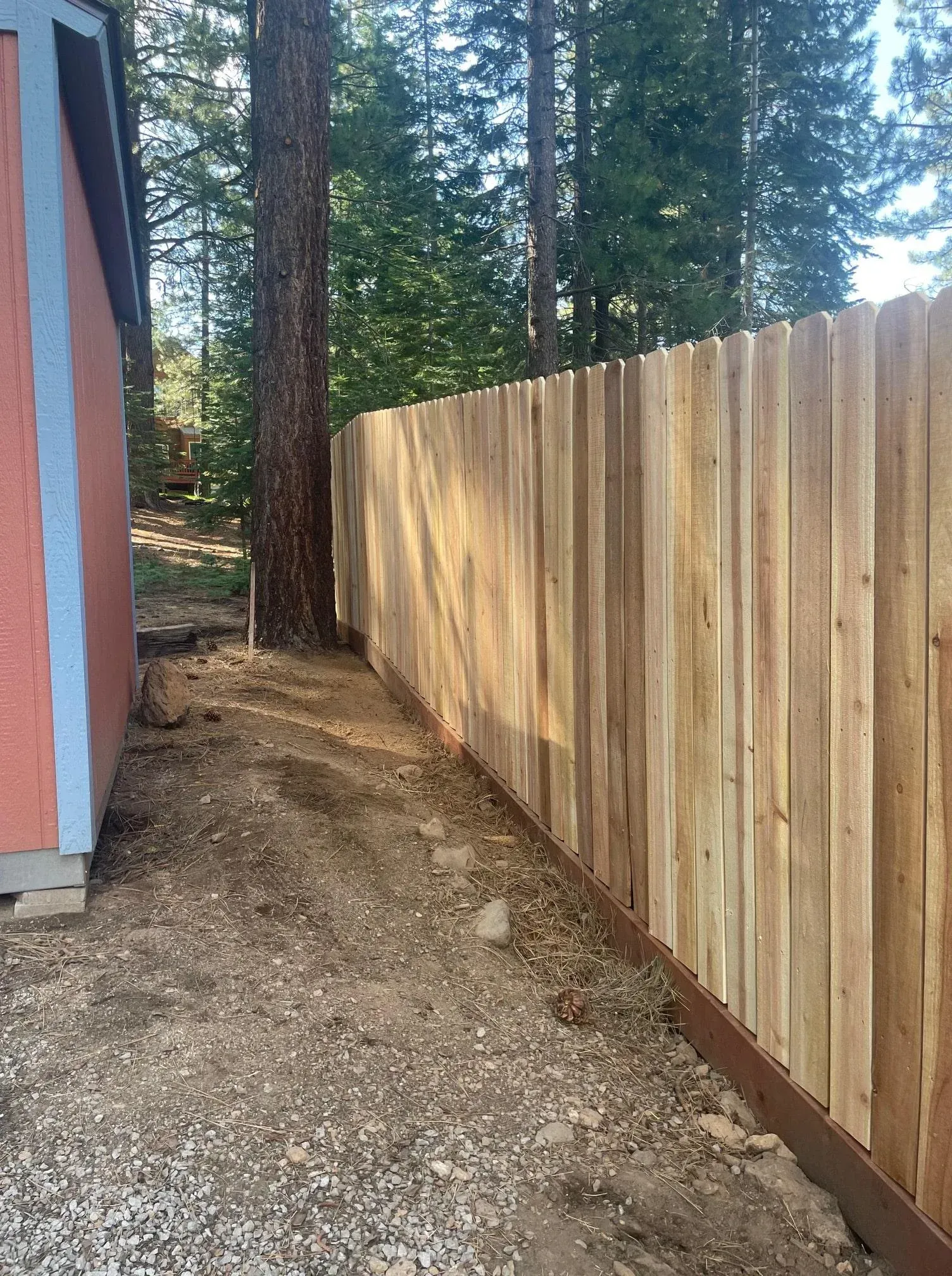 Wooden fence next to a building and trees on a gravel path, sunny day.